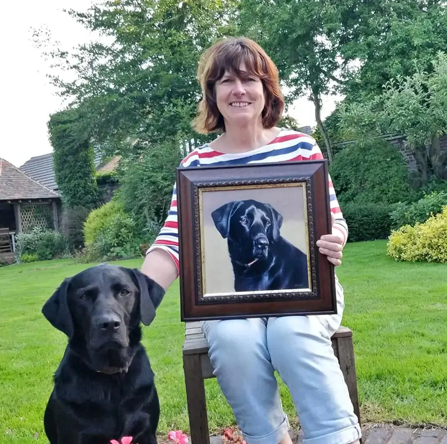 Mia the black Labrador proudly sitting with her framed birthday portrait and owner