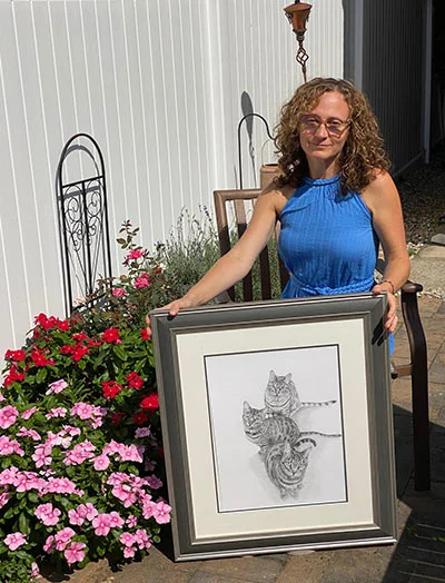 Erika smiling, holding two pet portraits of her cats, Lucy, Leo, and the newly adopted Lily, despite bright sunlight.